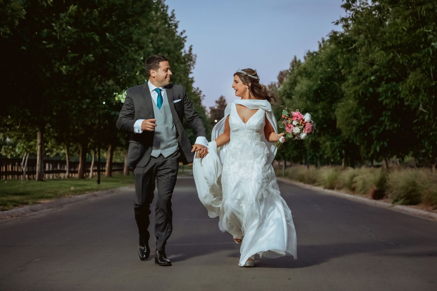 Bride and groom walking hand-in-hand down a tree-lined road on their wedding day.