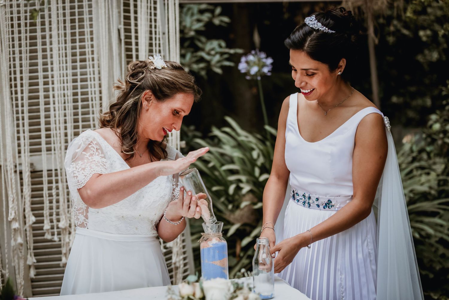 Two brides smile while performing a sand unity ceremony at their outdoor wedding.