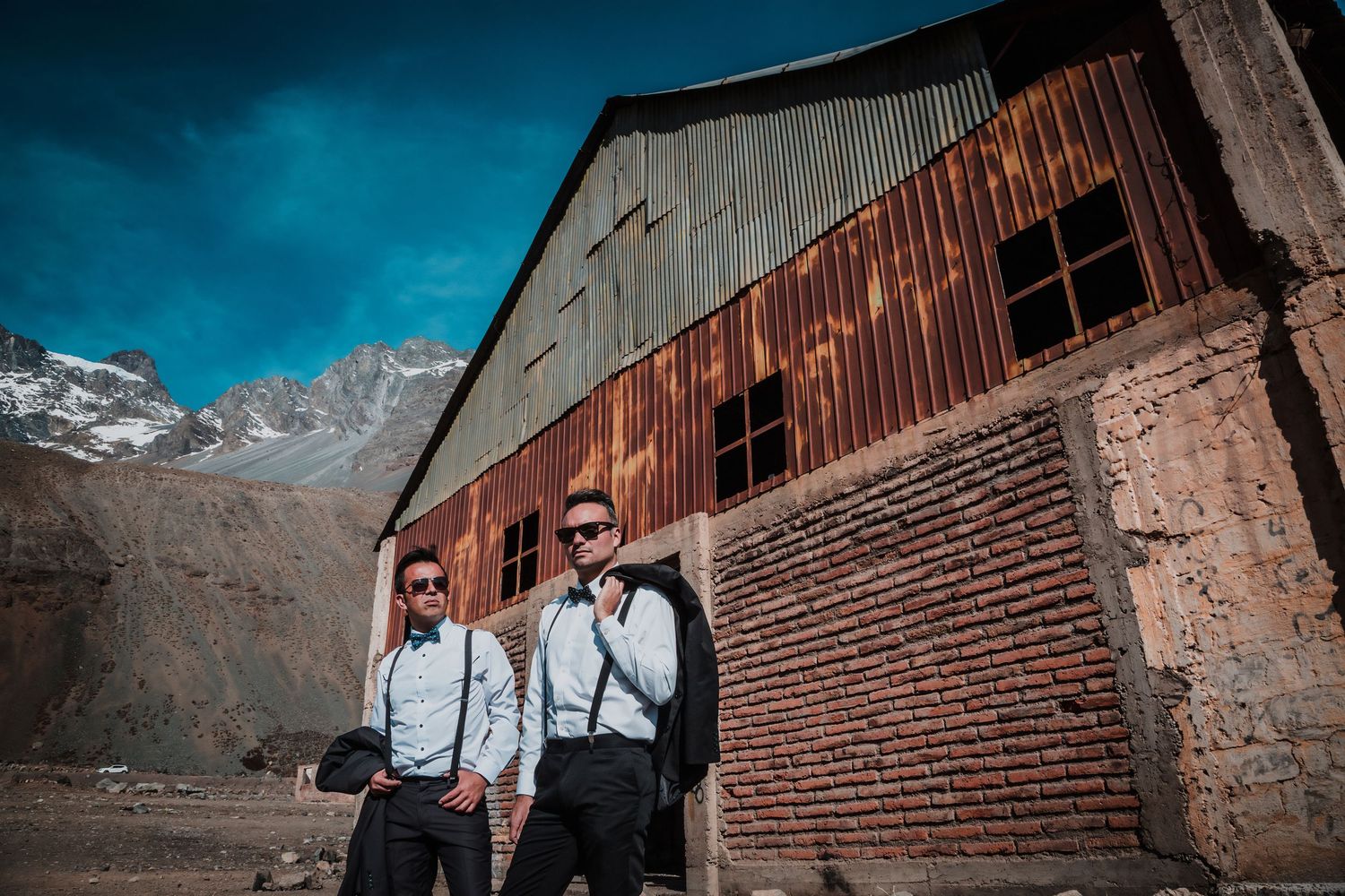 Two grooms in formal attire with suspenders pose confidently beside a weathered rustic barn and mountain scenery.