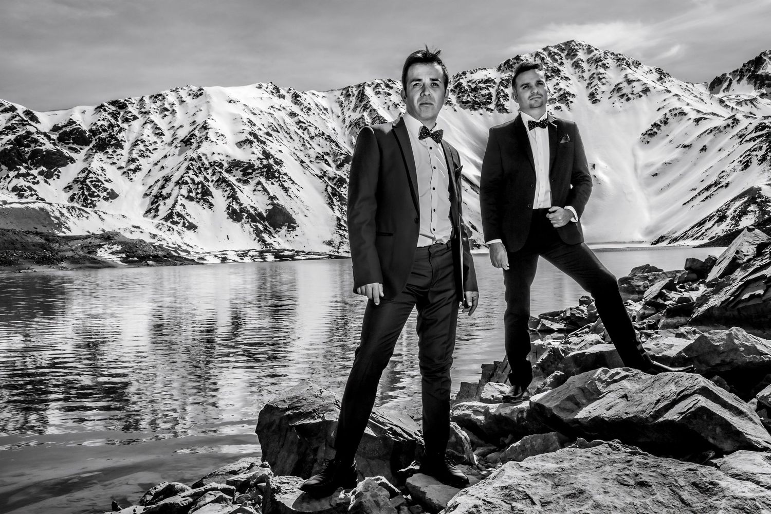 Black and white photo of two grooms in tuxedos posing by a glacial mountain lake.