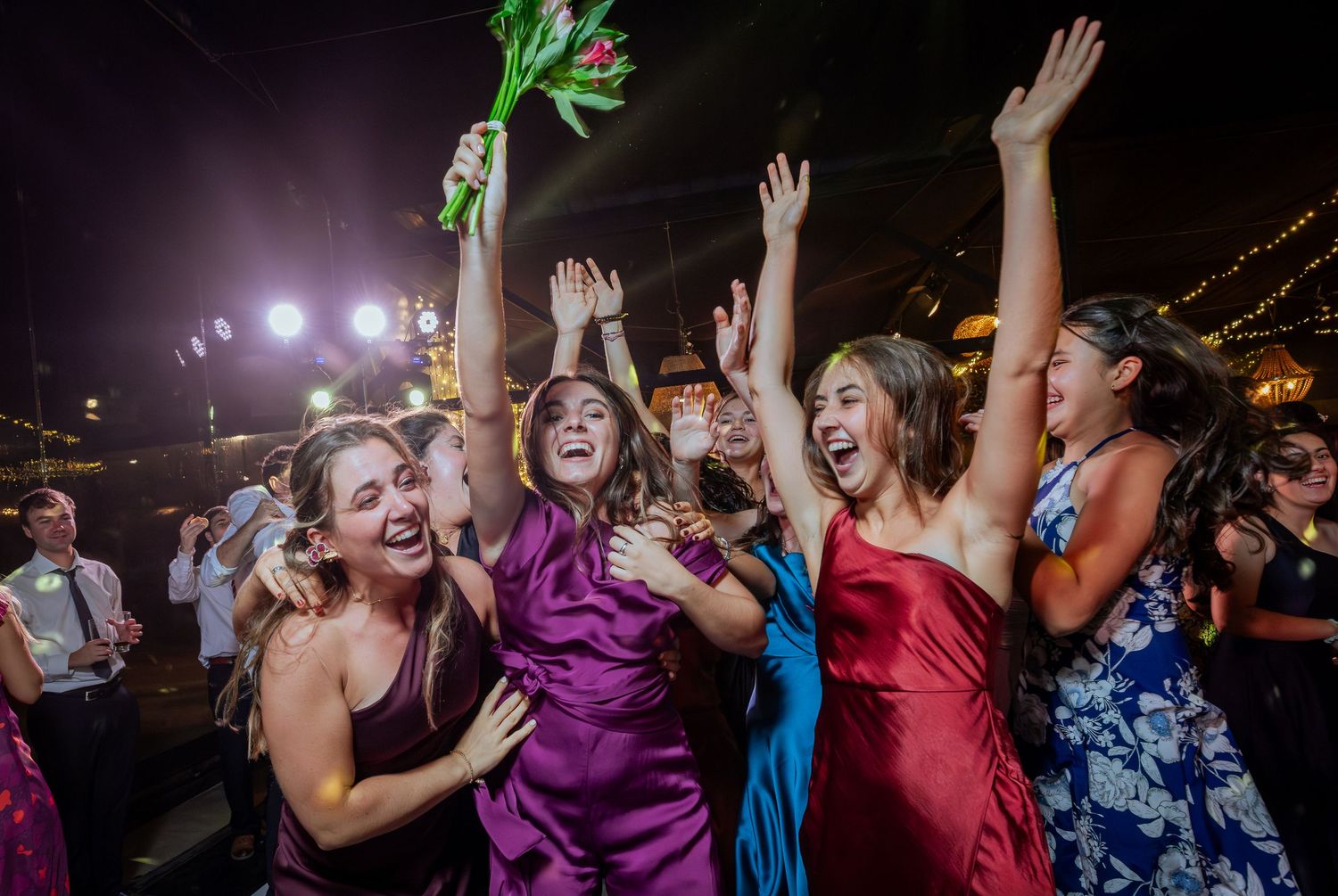 Excited bridesmaids in colorful dresses celebrate bouquet toss at wedding reception.