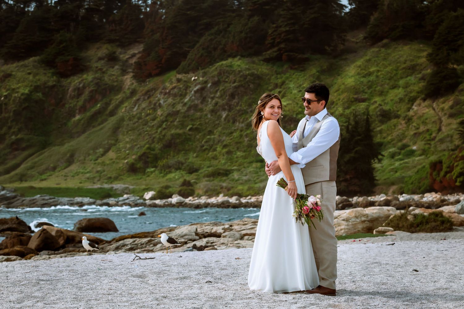 Bride and groom embracing on rocky beach with turquoise water and green cliffs in the background.
