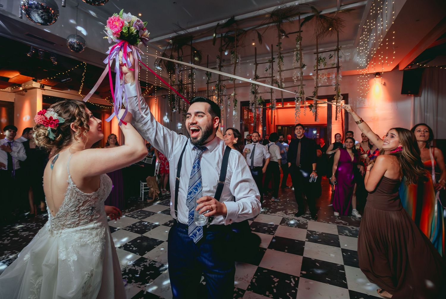 Excited groom holds up bridal bouquet with bride on confetti-filled wedding dance floor.