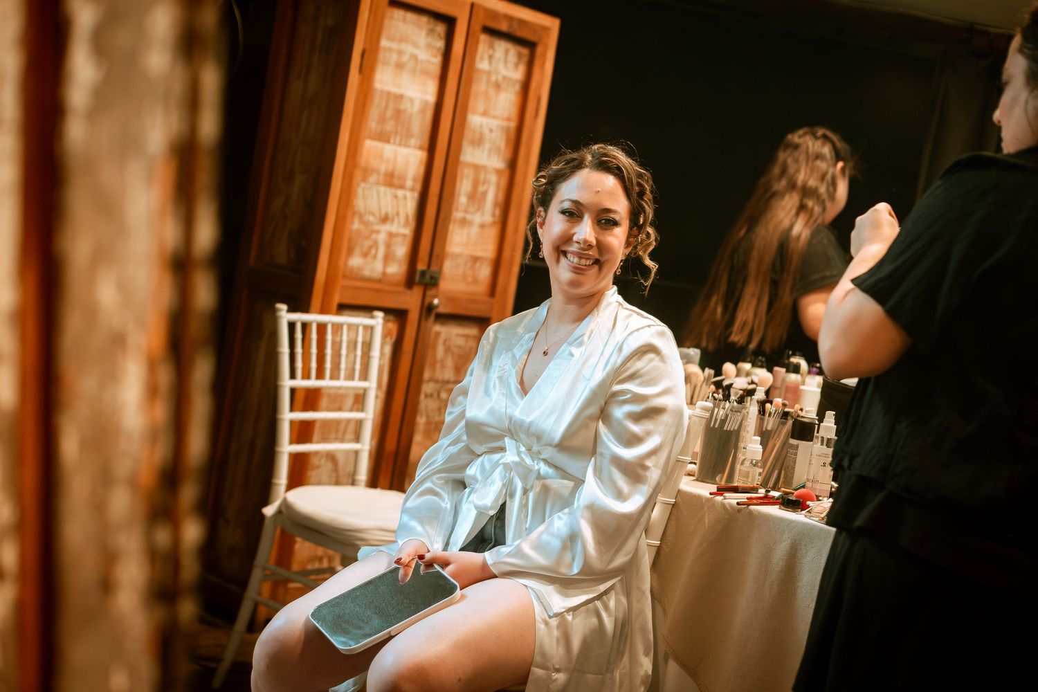 Smiling bride in white satin robe sitting during wedding preparations in elegant venue room.