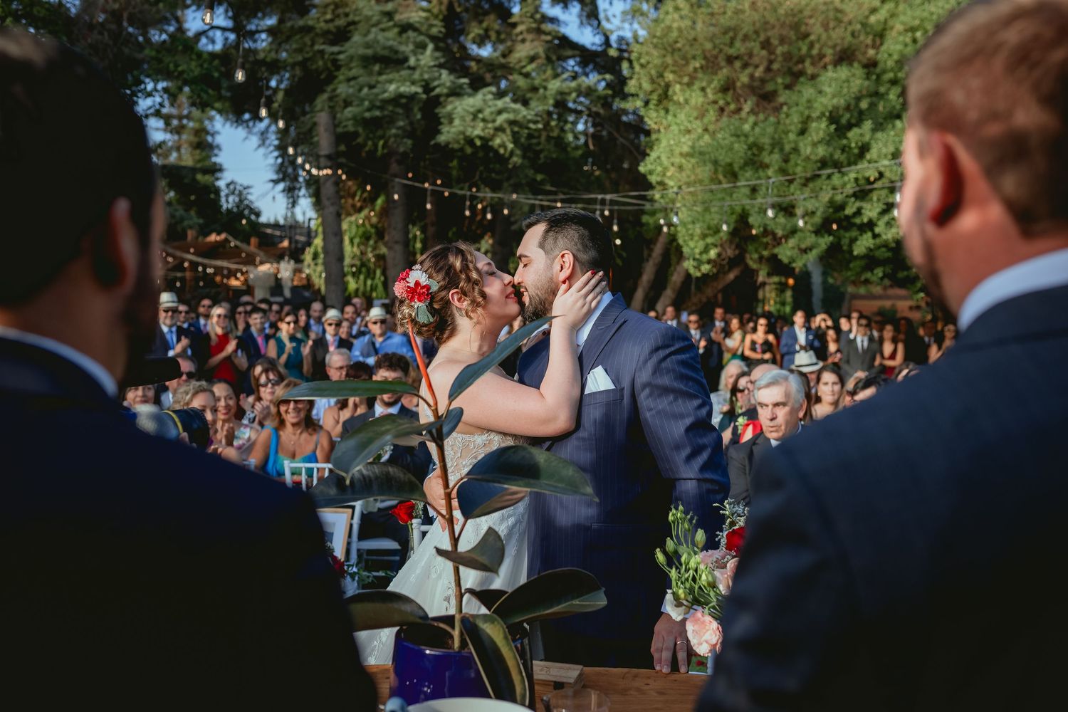 Bride and groom share first kiss at outdoor wedding ceremony with guests seated among trees and string lights.