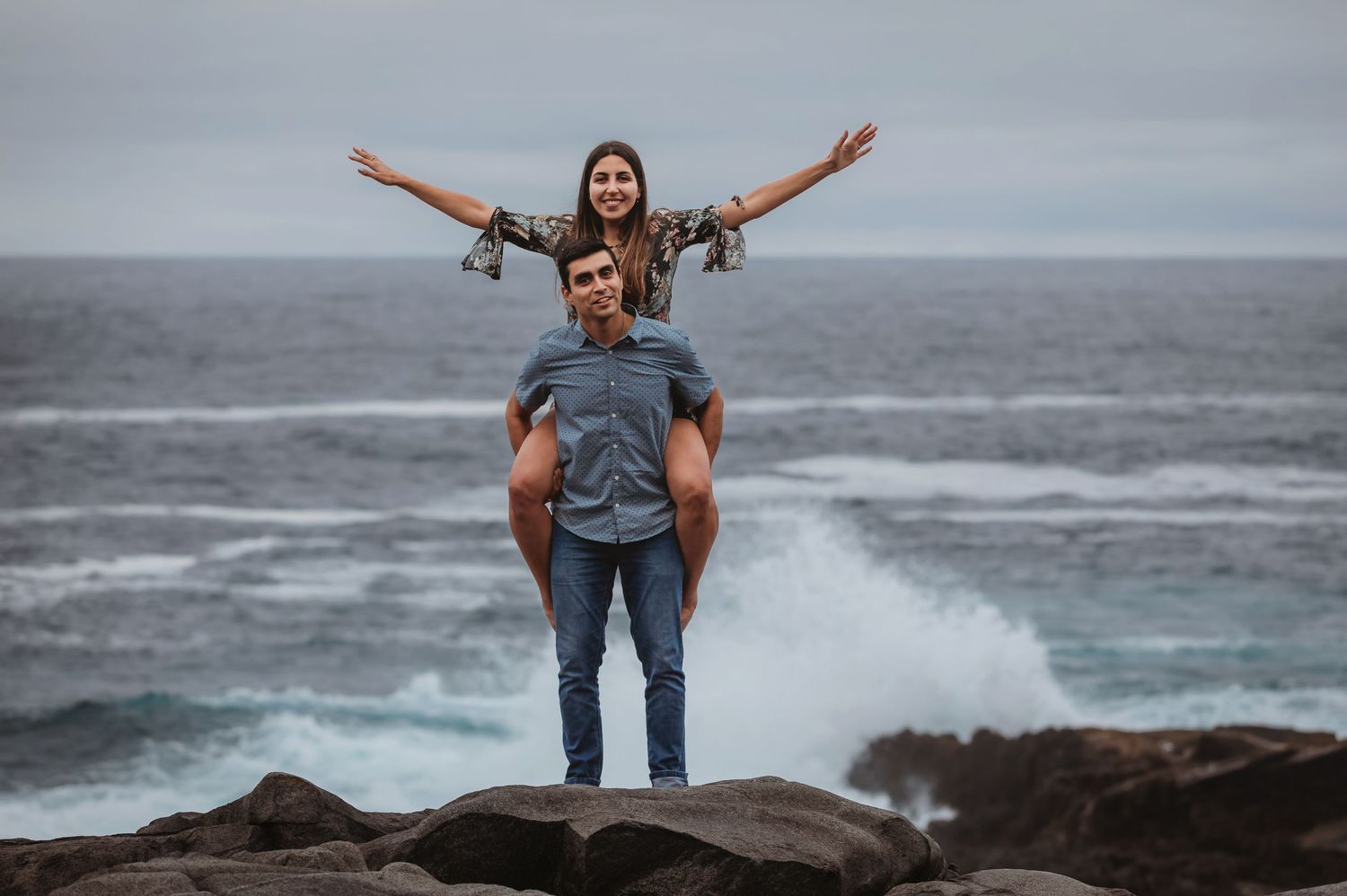 Woman sitting on mans shoulders with arms outstretched on rocky shore with crashing ocean waves behind them.