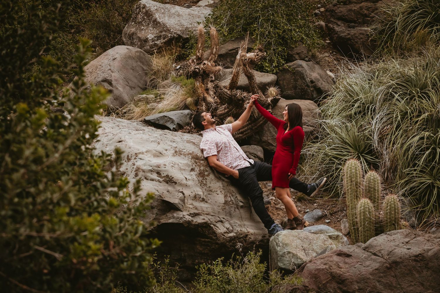 Couple playfully climbing rocks surrounded by cacti and desert vegetation outdoors.