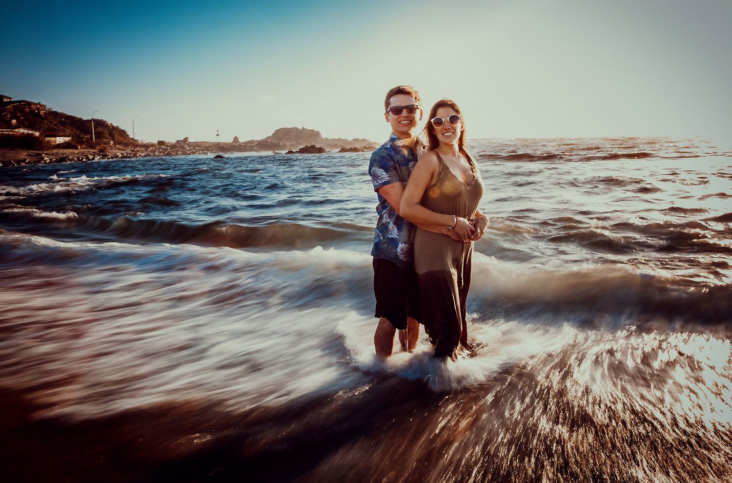 Couple standing together in shallow ocean waves on a bright sunny beach day.