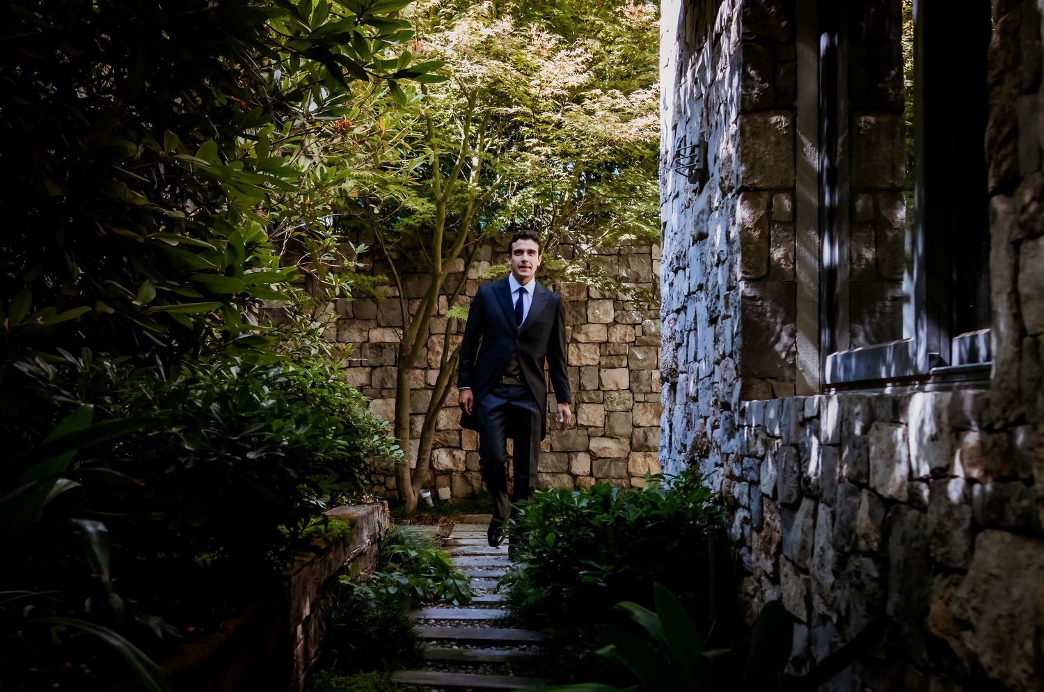 Groom in black suit walking up stone garden path surrounded by lush green foliage.