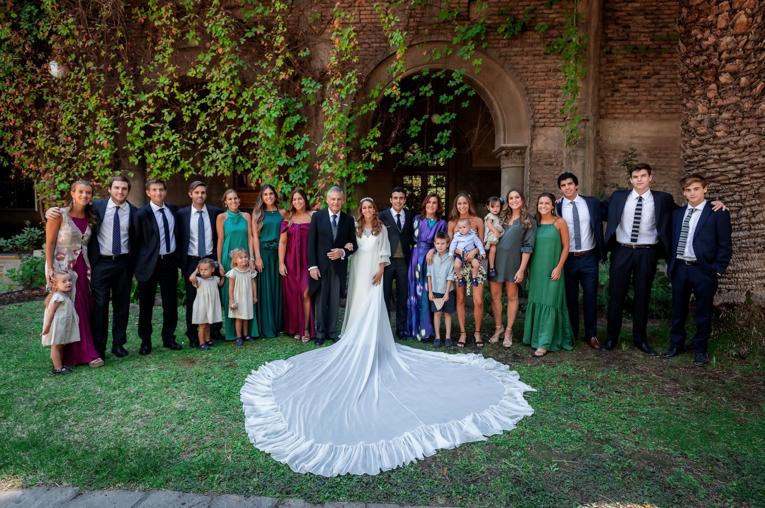 Bride in long cathedral veil poses with large wedding party outdoors at historic venue.