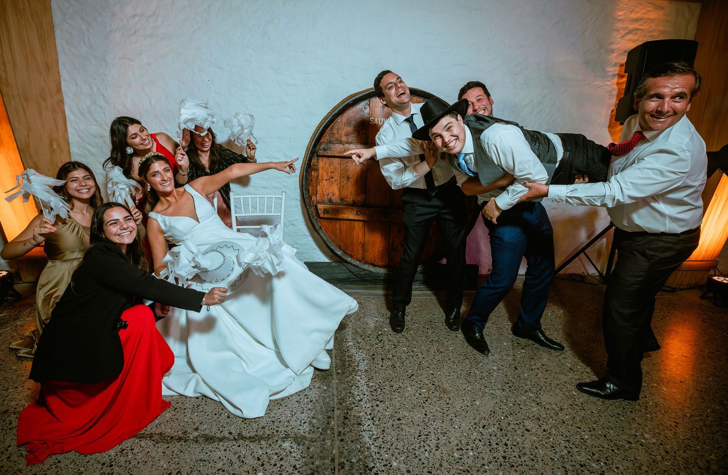 Wedding party poses playfully with bride and groom near rustic barrel at reception venue.