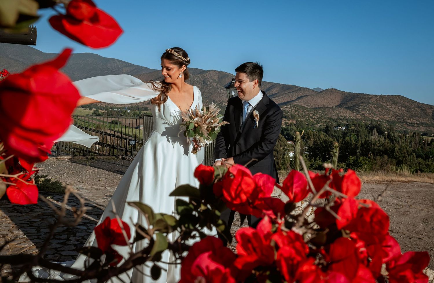 Bride and groom walking hand in hand near red bougainvillea with scenic mountain views.