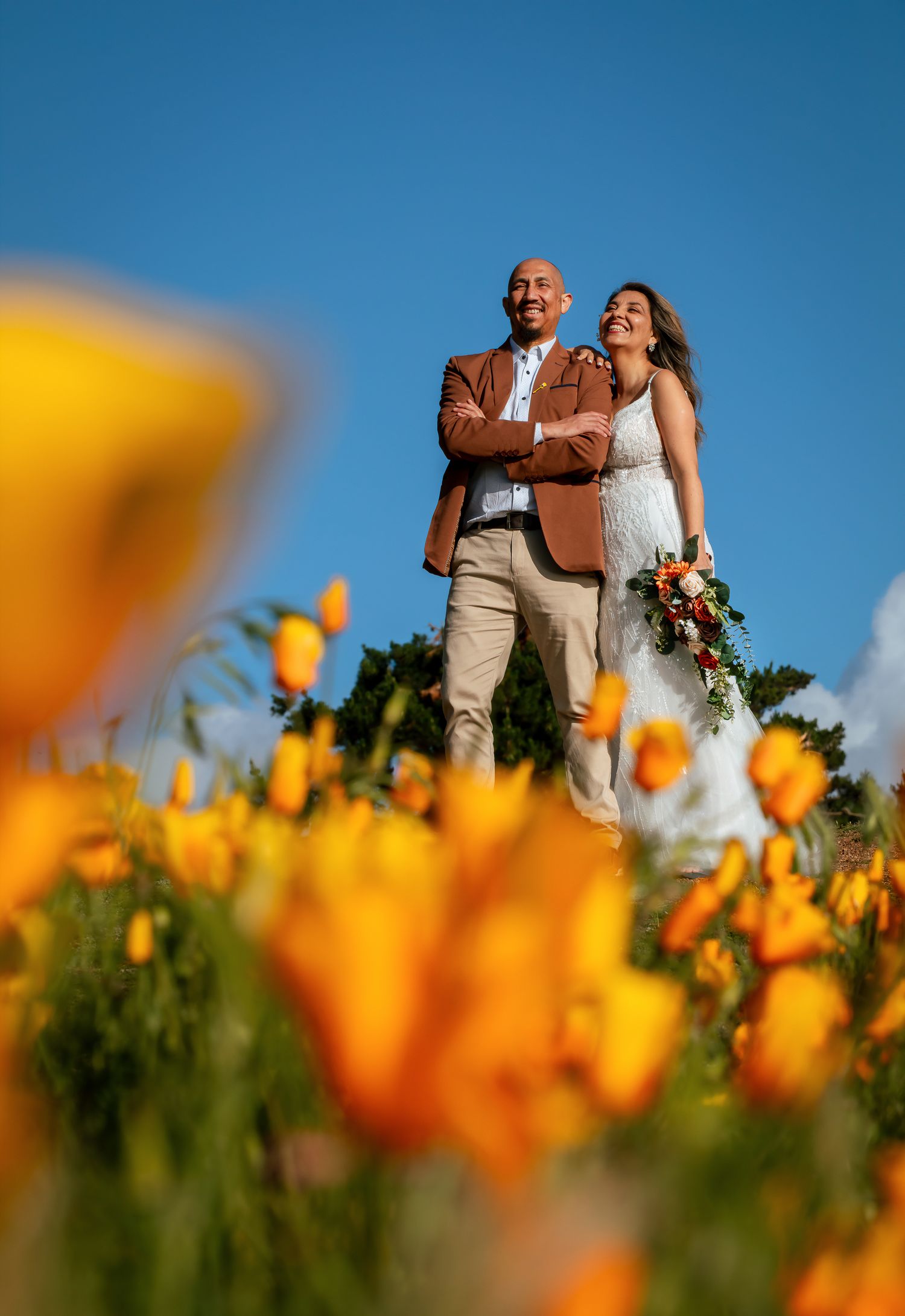 Smiling couple in wedding attire poses arm in arm above a field of bright orange poppies under a clear blue sky.
