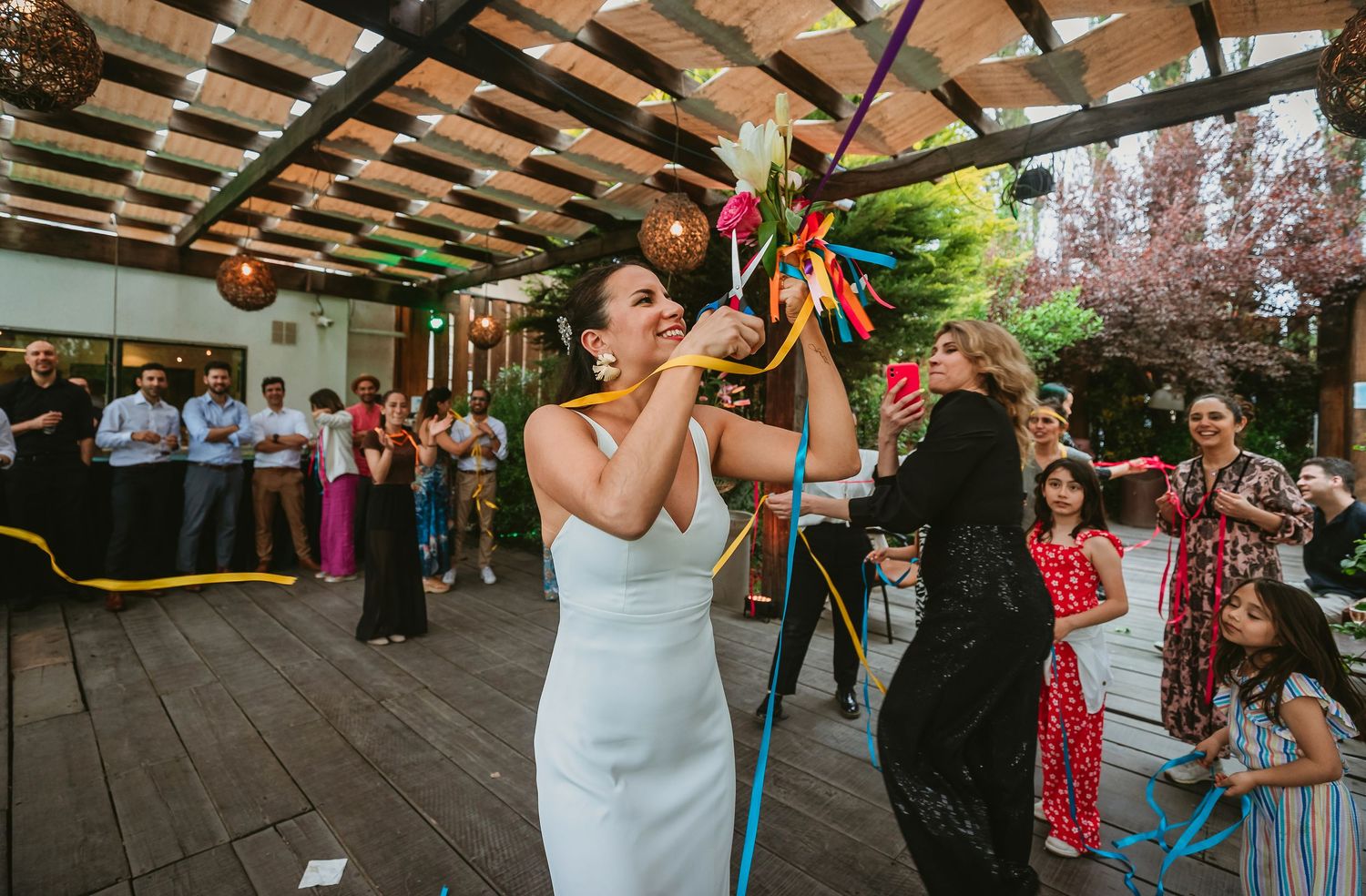 Bride in white dress breaking a colorful pinata at an outdoor wedding reception celebration.