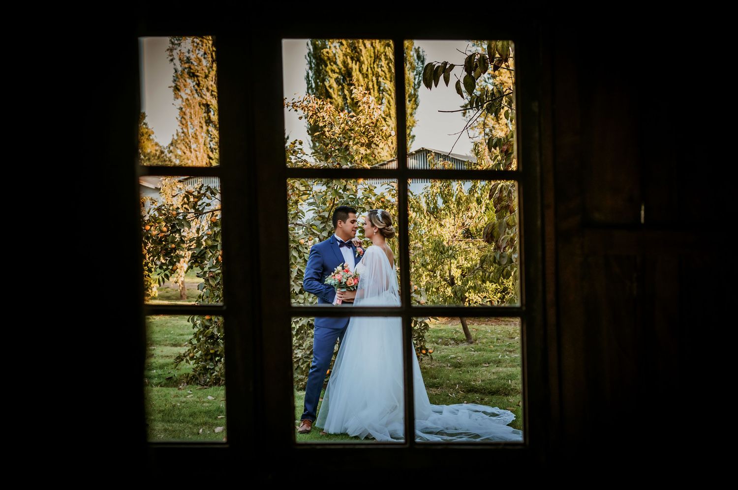 Bride and groom kissing in a garden, viewed through a rustic window frame on their wedding day.