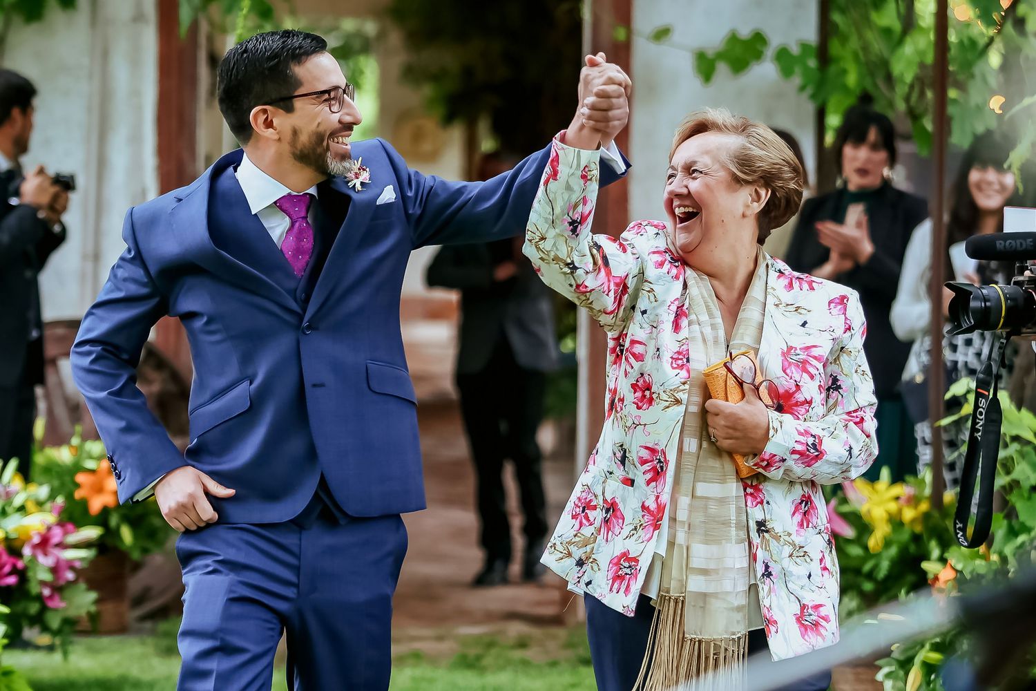 Joyful groom in blue suit celebrating with laughing woman in floral jacket at outdoor wedding.
