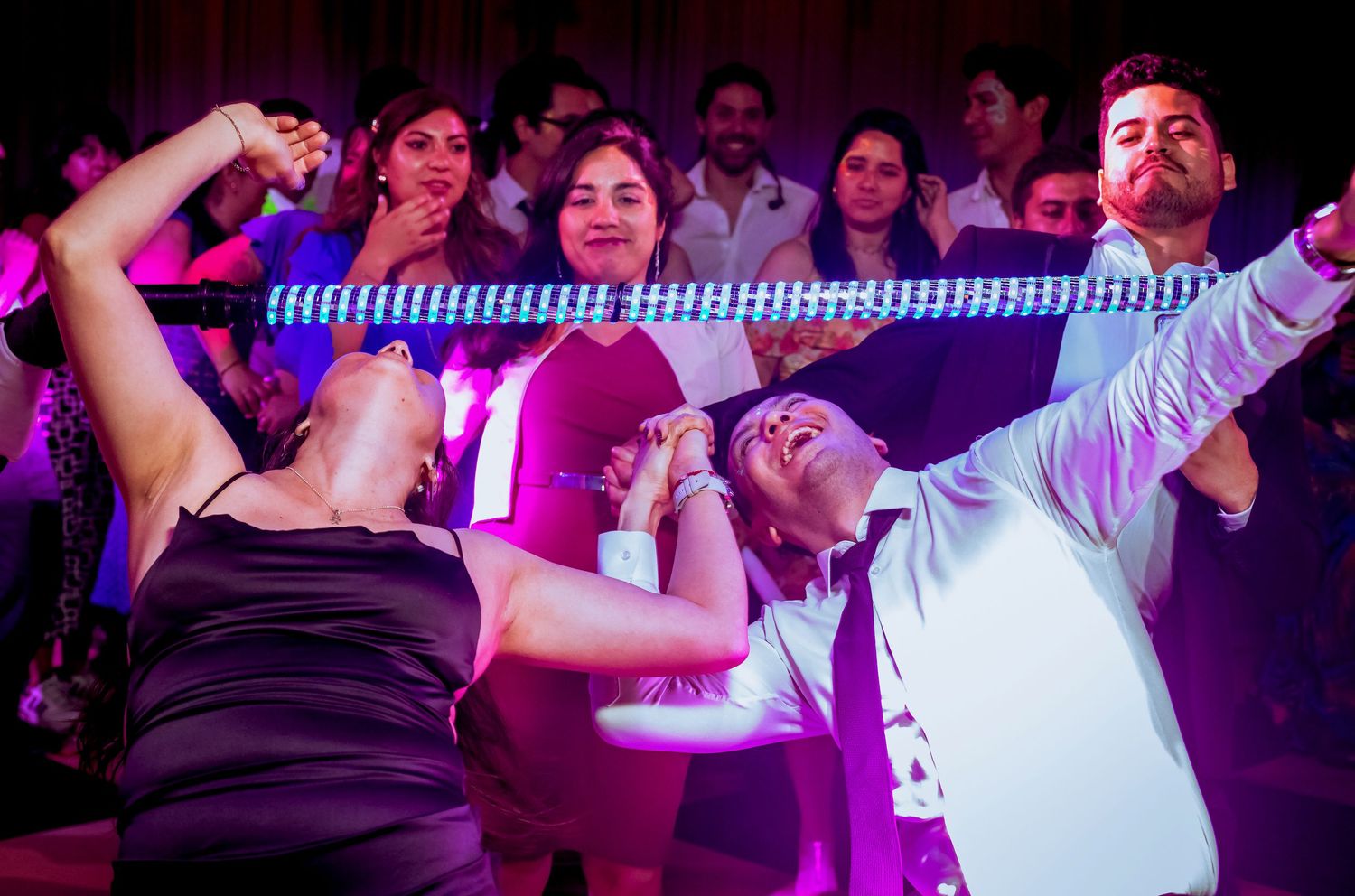 Guests laugh and dance under a glowing LED limbo bar at a lively wedding reception.