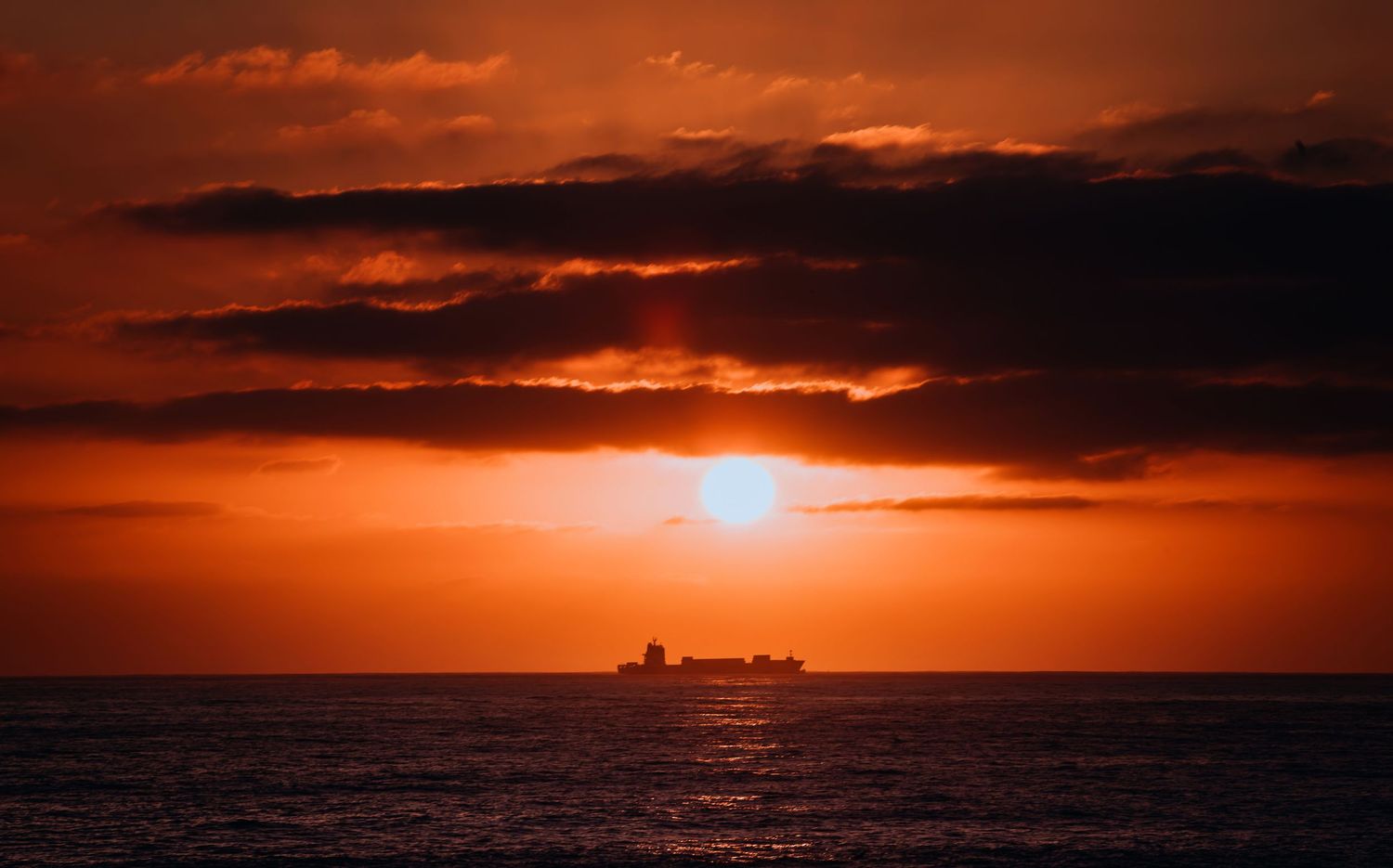 A cargo ship silhouetted against a vibrant red and orange sunset over the calm ocean horizon.