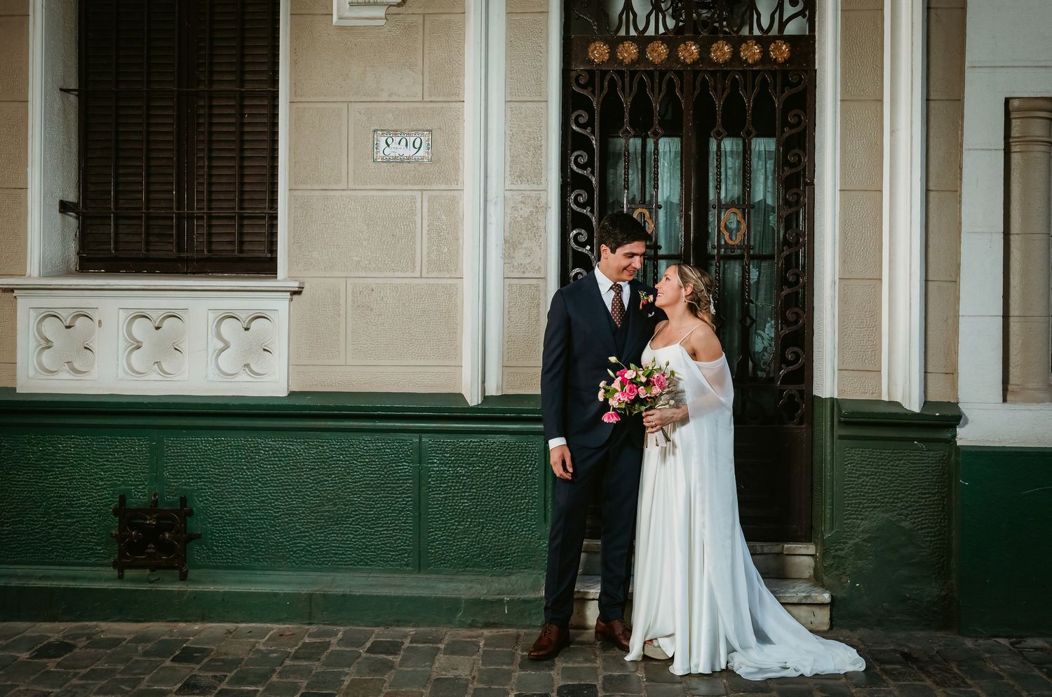 Bride and groom share romantic glance outside charming green and white building.