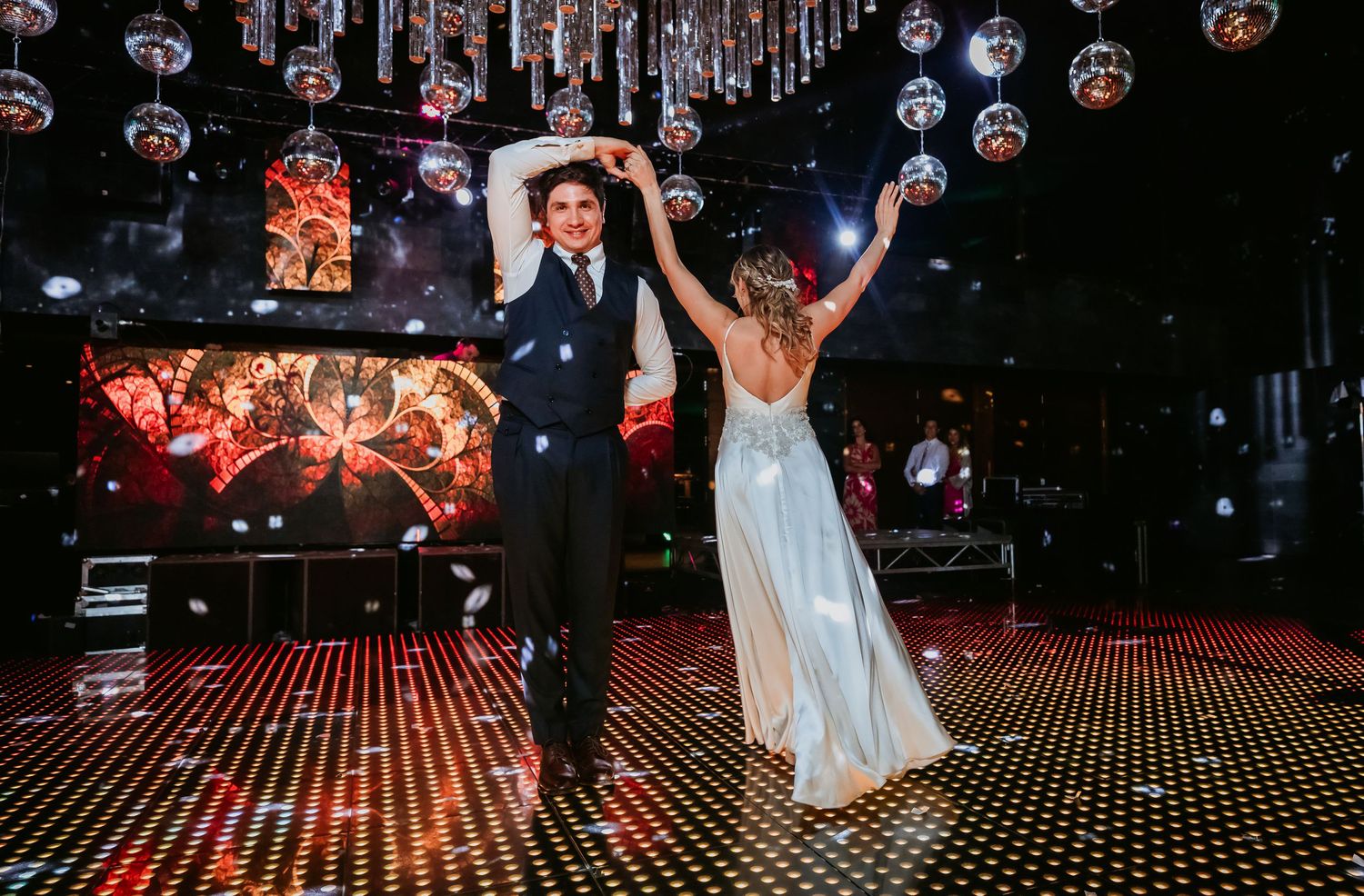 Groom spins bride in wedding dress on glittering dance floor under disco balls at reception.