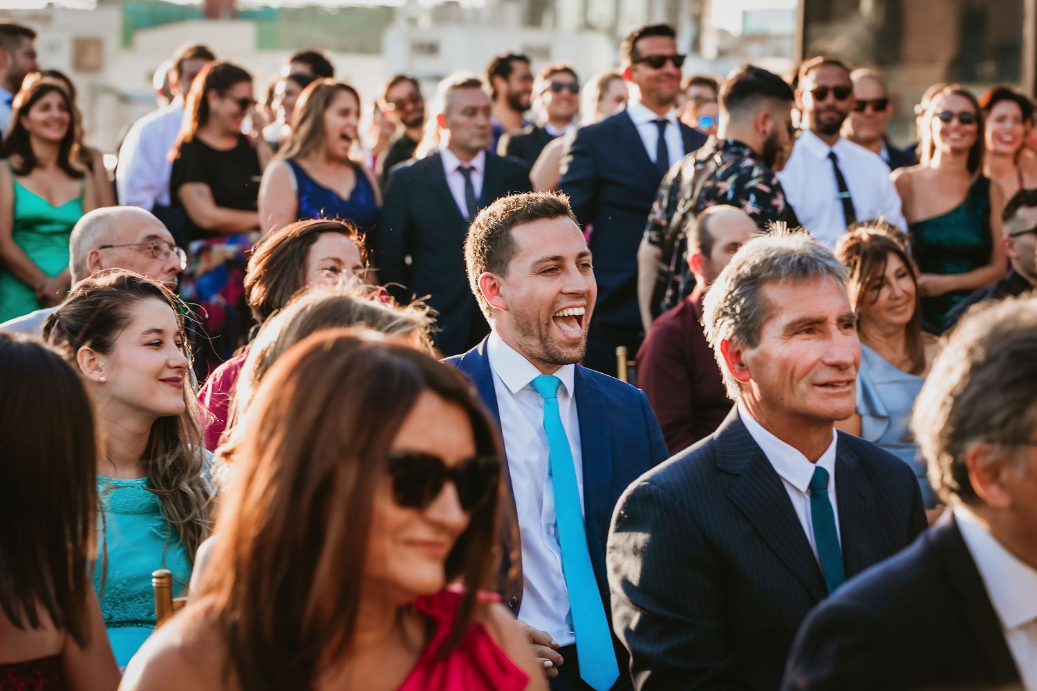 Smiling wedding guests in formal attire enjoying an outdoor sunny ceremony.