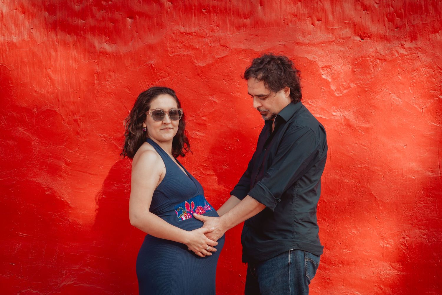 Couple tenderly holding pregnant belly against a vibrant red wall.