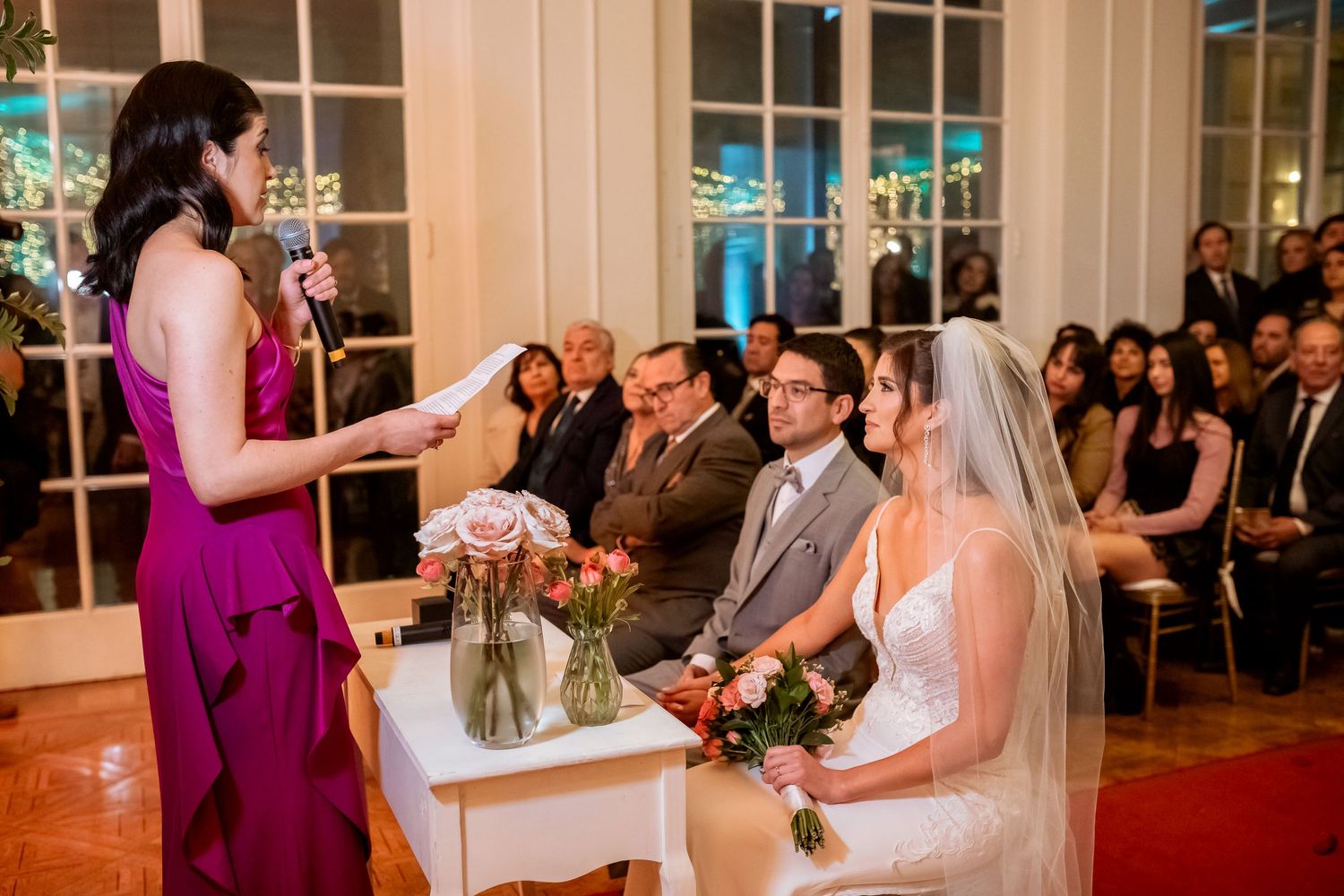 A bridesmaid in purple reads a speech at an elegant indoor wedding ceremony with the bride and groom seated at the altar.