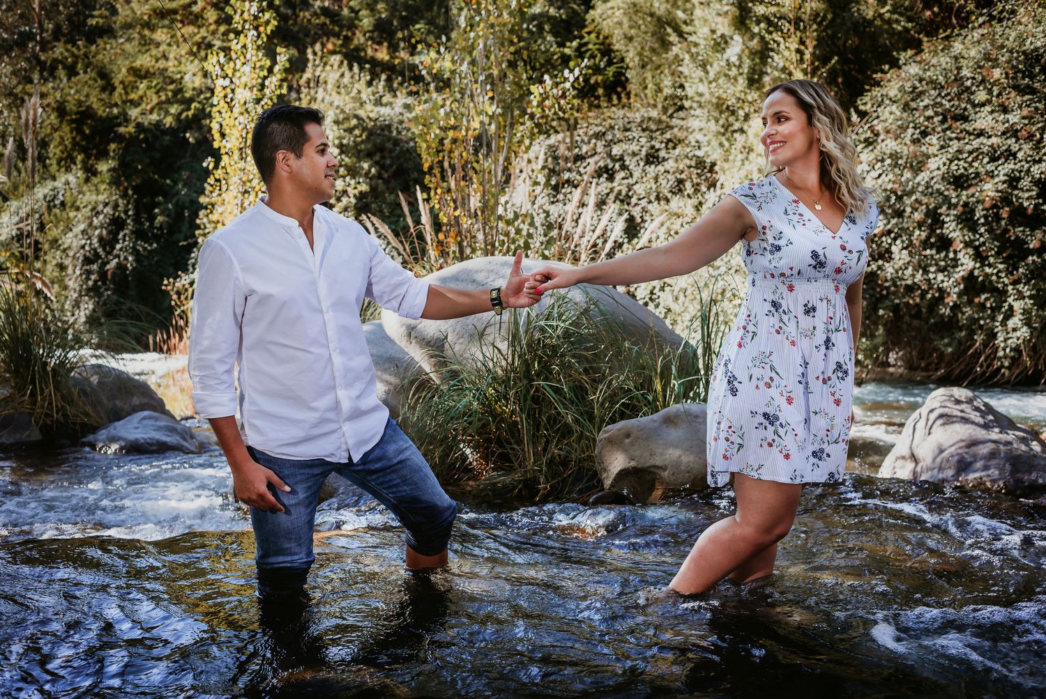 Couple holding hands in a rocky stream surrounded by lush greenery, man in white shirt and woman in floral dress.
