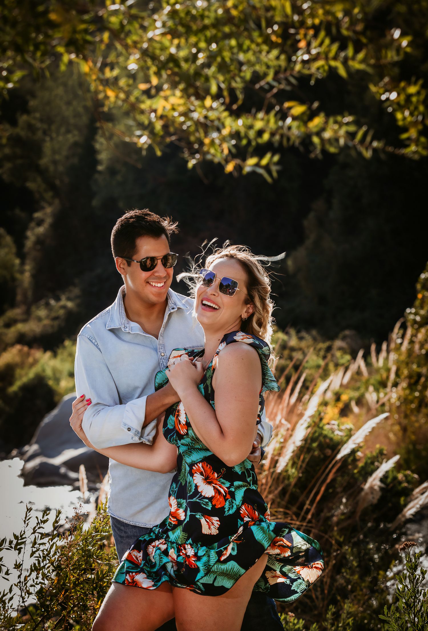 A smiling couple in sunglasses embracing outdoors, woman in floral dress, man in light blue shirt, with lush greenery behind them.