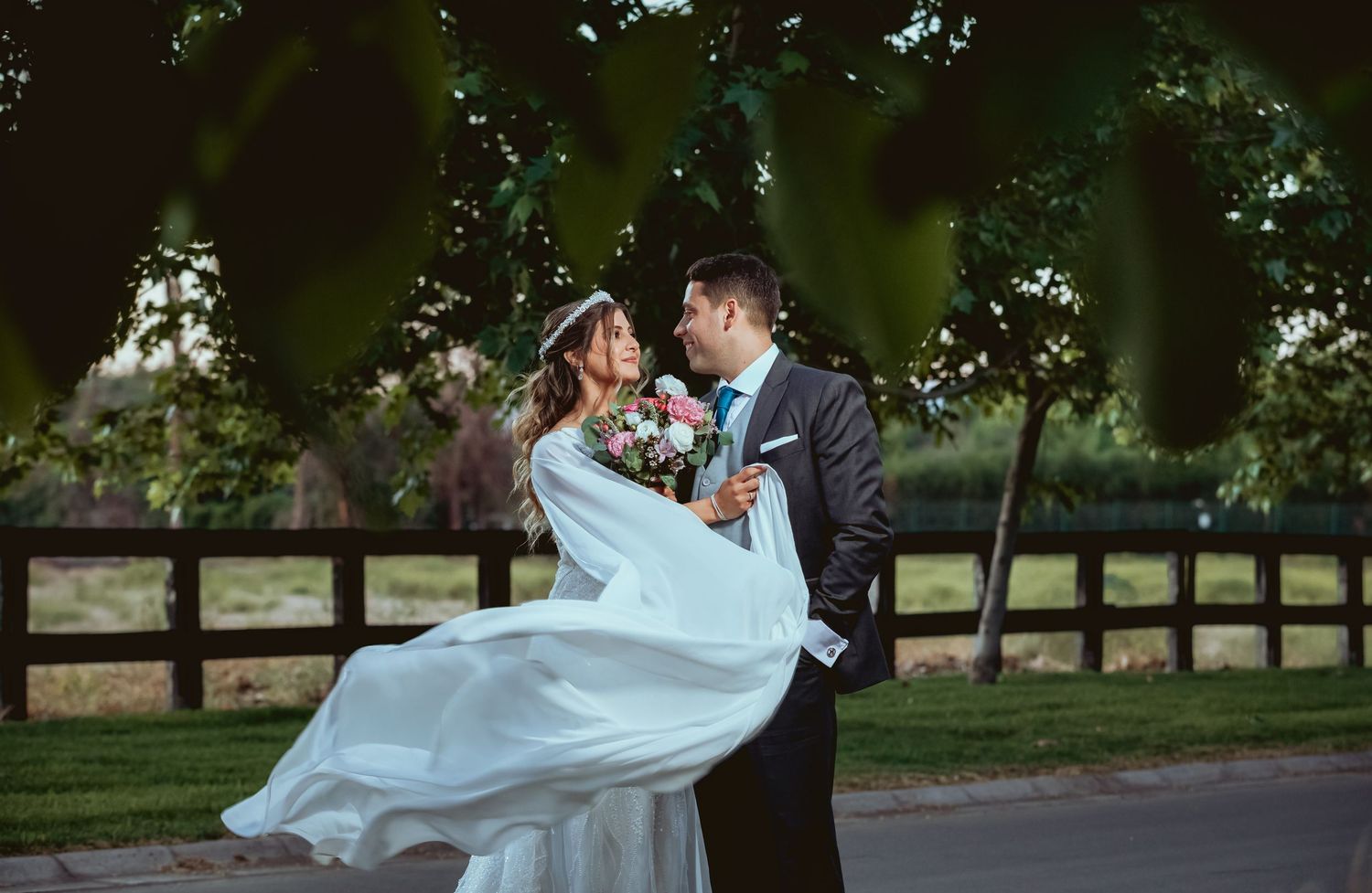 A groom lifts his bride in a white flowing gown outdoors near a wooden fence surrounded by lush green trees.