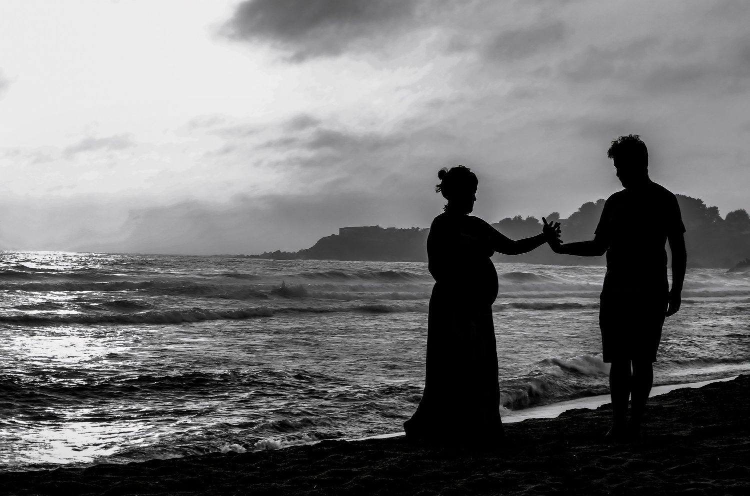 Silhouette of a couple standing on a beach at sunset, with ocean waves and dramatic cloudy sky in black and white.