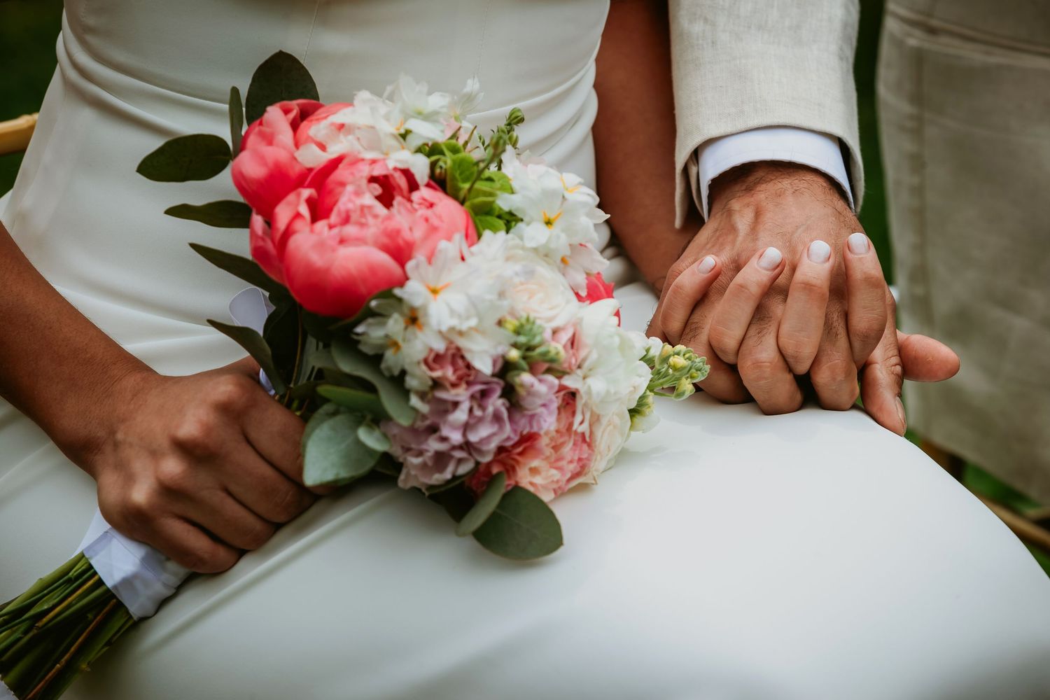 Bride and groom holding hands with a pink peony and white flower wedding bouquet on a white dress.