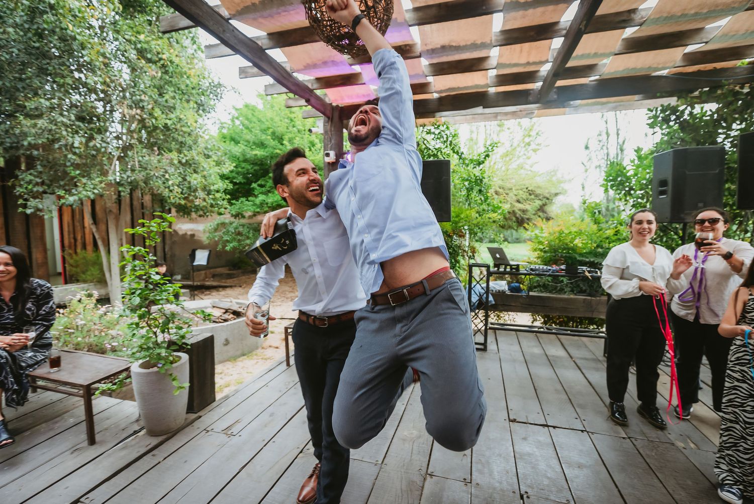 Two men jumping and celebrating on a wooden deck at an outdoor wedding reception party.