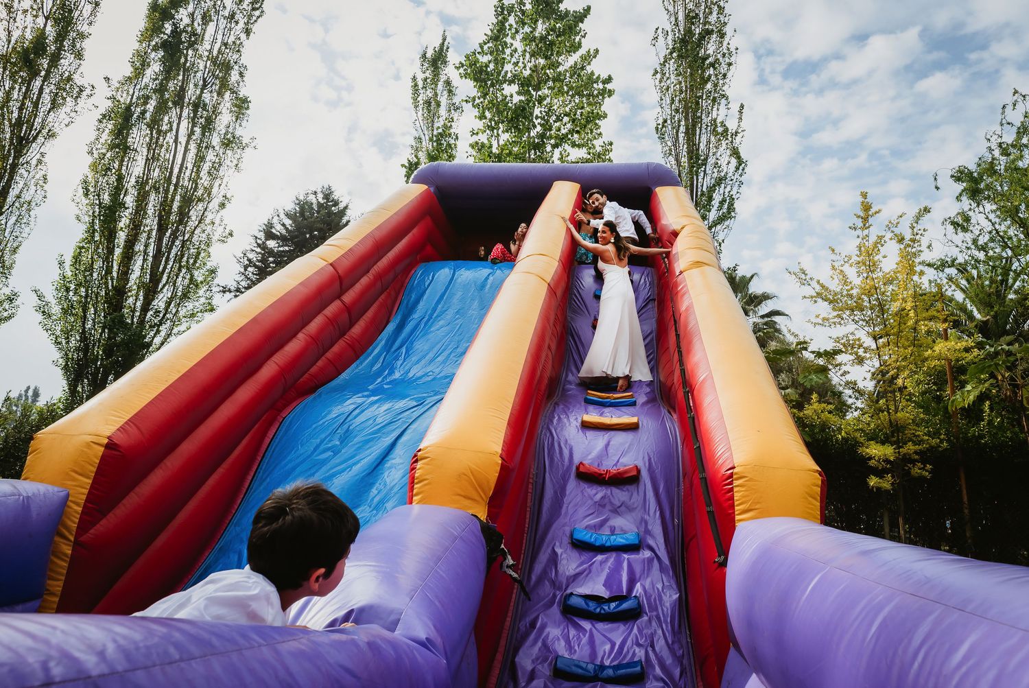 Children playing on a colorful inflatable bounce house slide outdoors surrounded by tall trees.