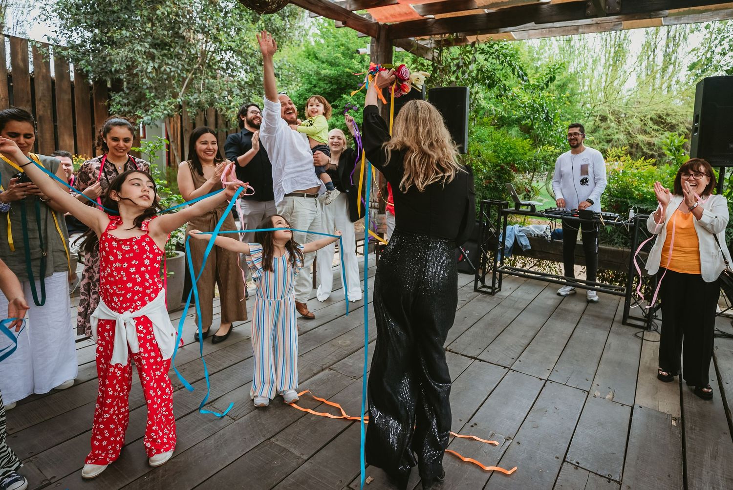 A lively outdoor party on a wooden deck with guests dancing and a performer in a black dress leading the celebration.
