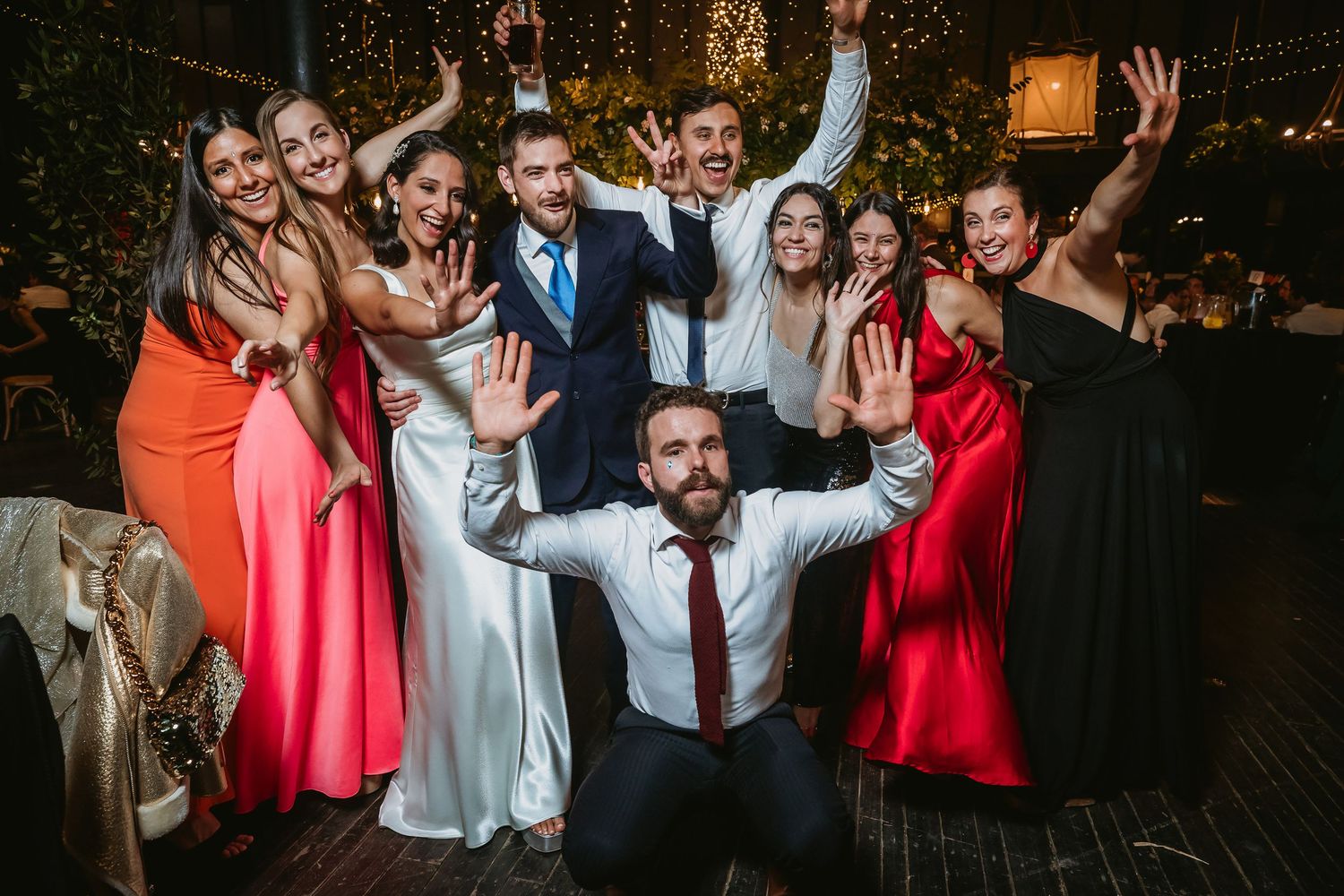 Wedding party group photo with bride, groom, and guests celebrating on dance floor with raised hands.