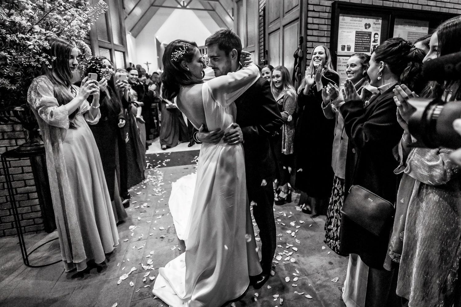 Black and white photo of newlyweds sharing a kiss while guests celebrate with confetti outside a church.