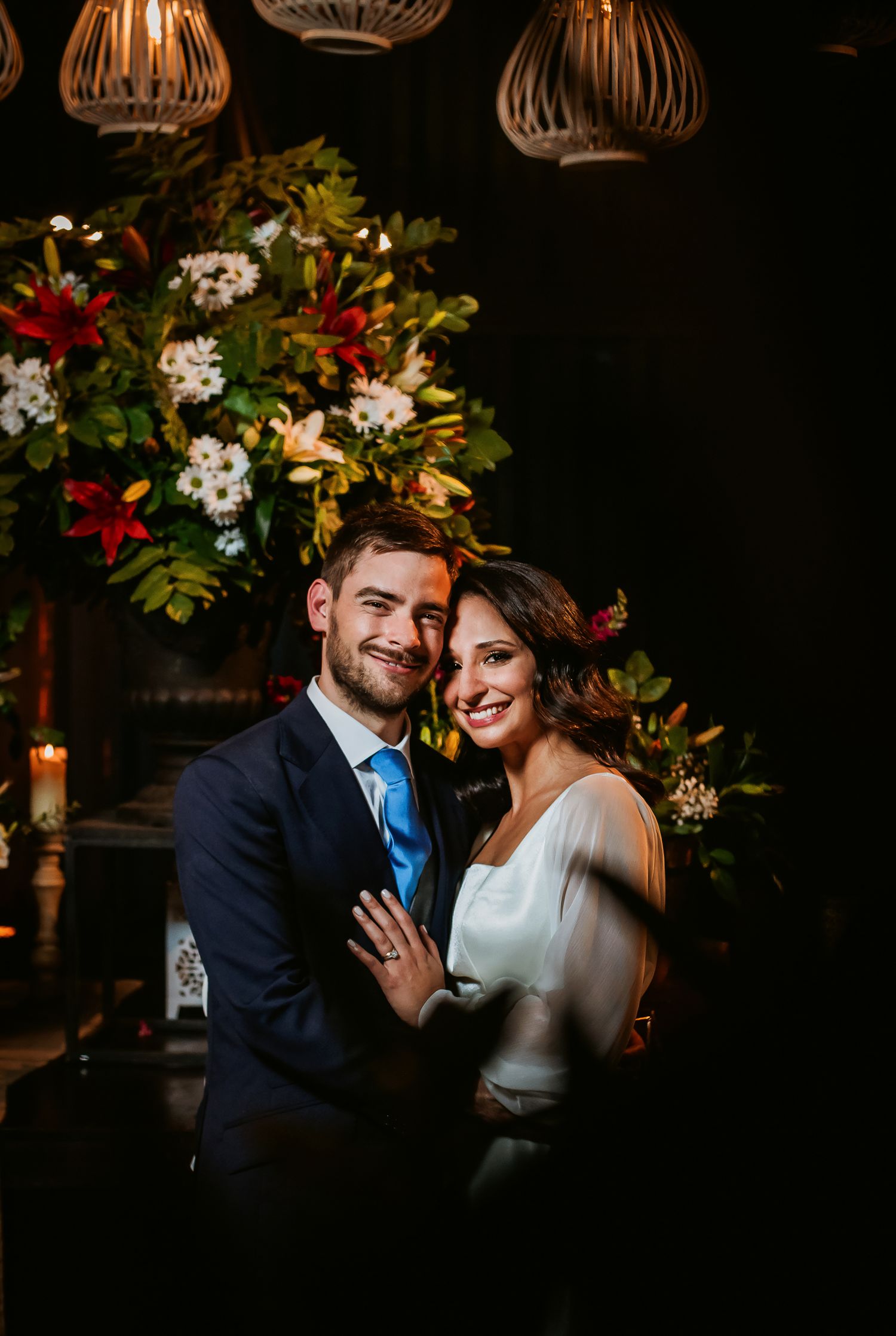 Bride and groom smiling together in front of lush floral arrangement with warm candlelight at their wedding reception.