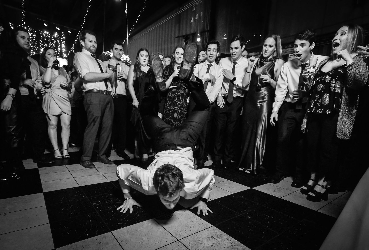 Black and white photo of a man breakdancing on the floor at a wedding reception while guests cheer and watch.