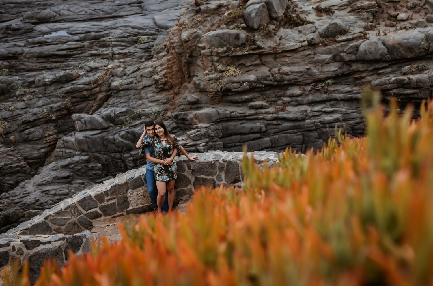 Couple walking along stone path with vibrant orange succulent plants and rocky cliffs.