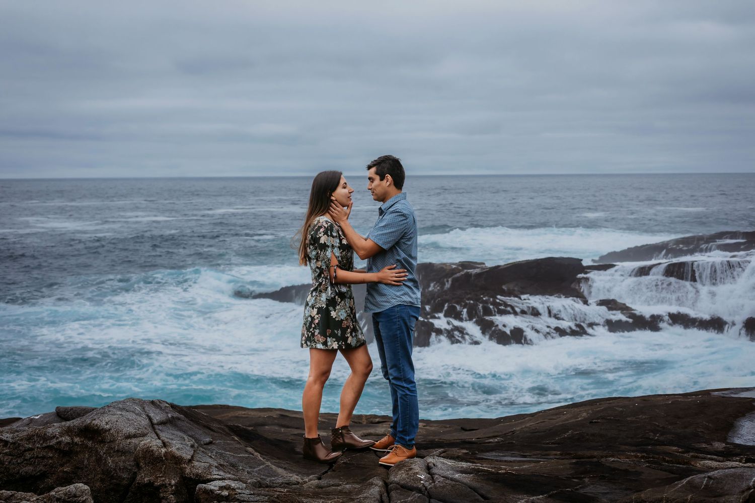 Couple embracing on rocky coastline with dramatic blue ocean waves splashing in background.