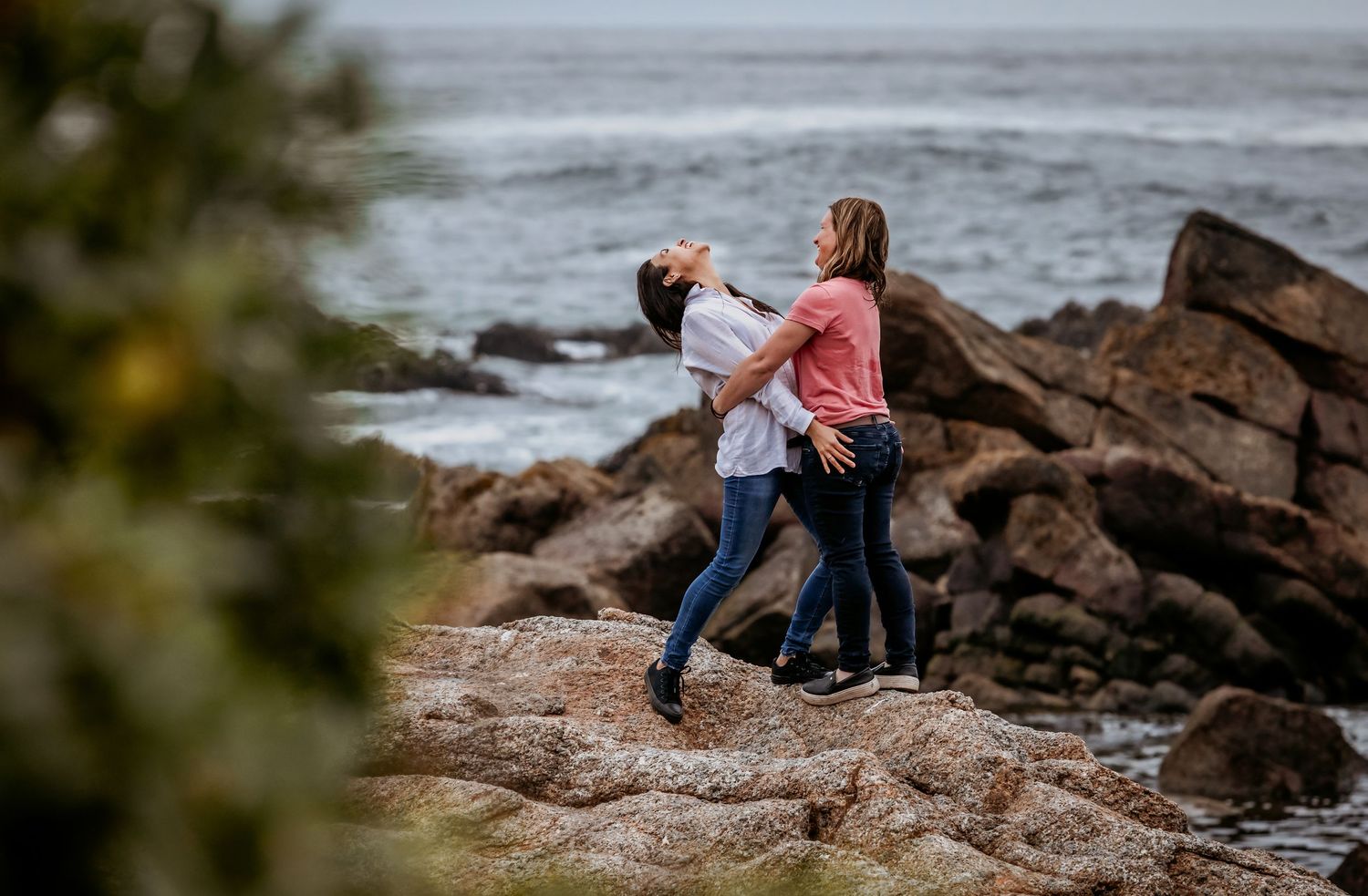 Two women laugh and dance together on rocky coastal boulders by the sea.