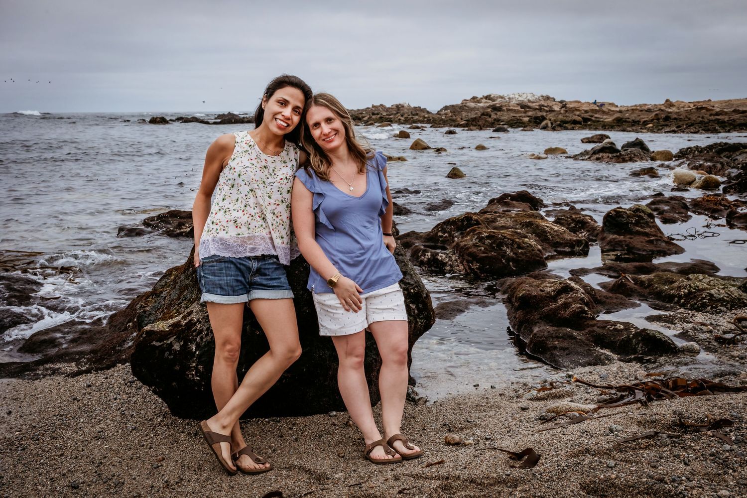 Two smiling women pose together leaning against a rock on a scenic rocky beach.