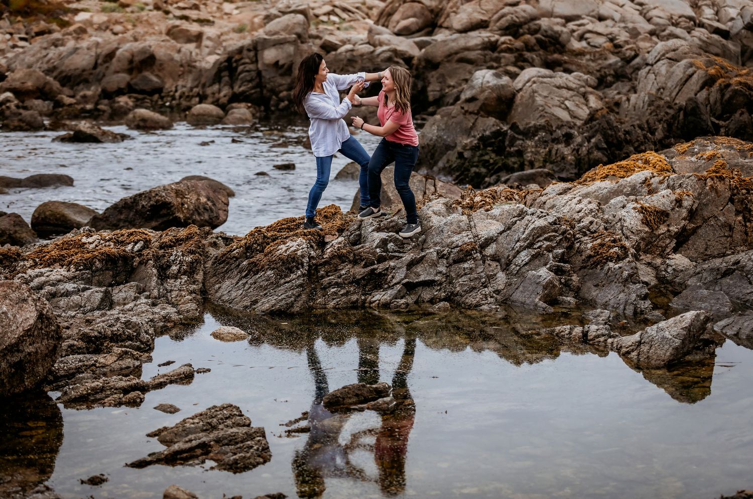 Two women dance playfully on rocks with their reflection visible in tide pools.