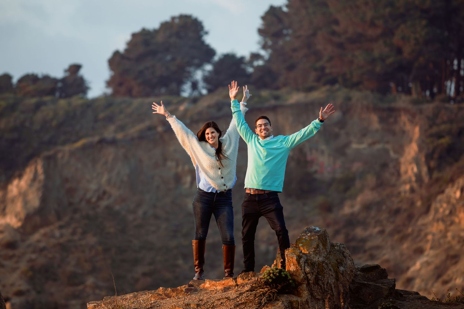 Joyful couple raising arms in celebration on rocky coastal bluff with tree-lined cliffs behind them.