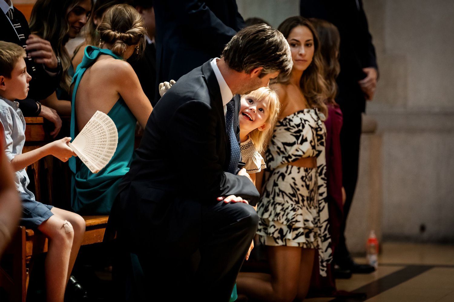 Wedding guests laughing and chatting in church pews during a ceremony, man in suit kneeling beside seated guests.