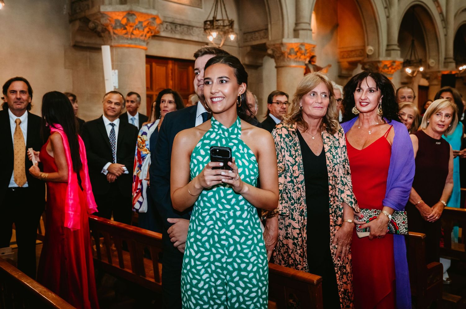 Wedding guests in colorful attire standing in church pews, woman in green dress holding phone.