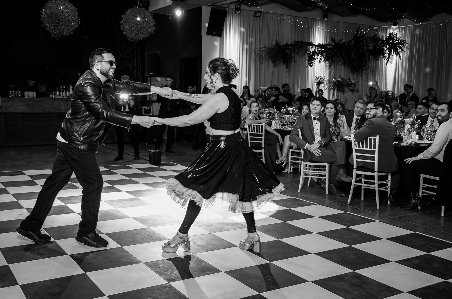 Couple performs energetic swing dance on checkered floor at elegant wedding reception in black and white photo.