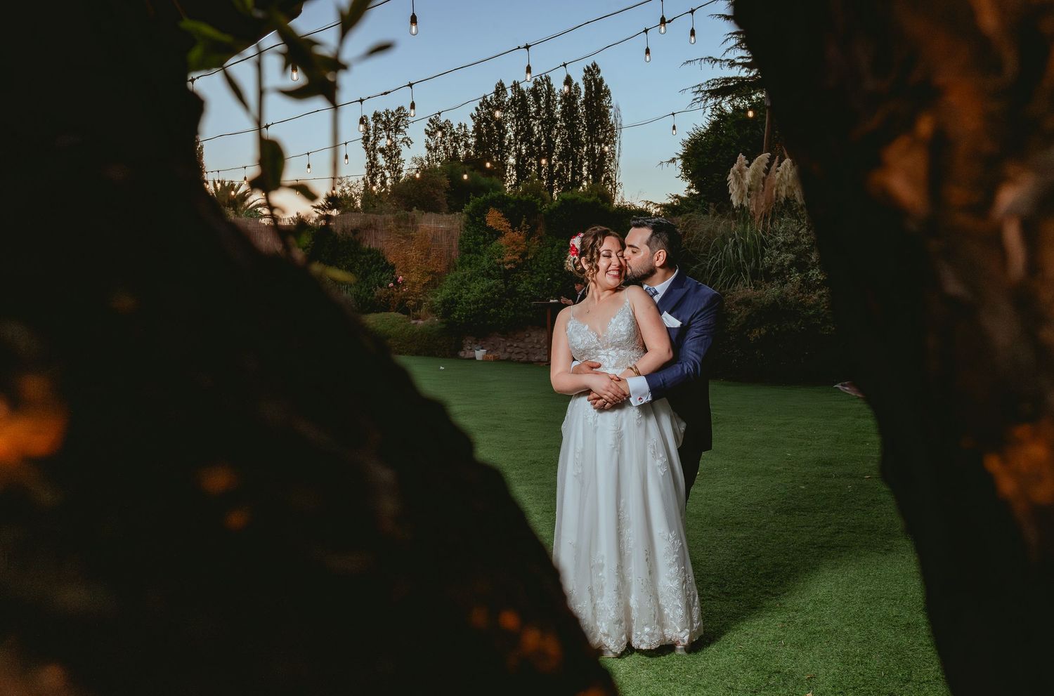 Wedding couple embraces tenderly in lush garden at golden hour framed by tree branches.