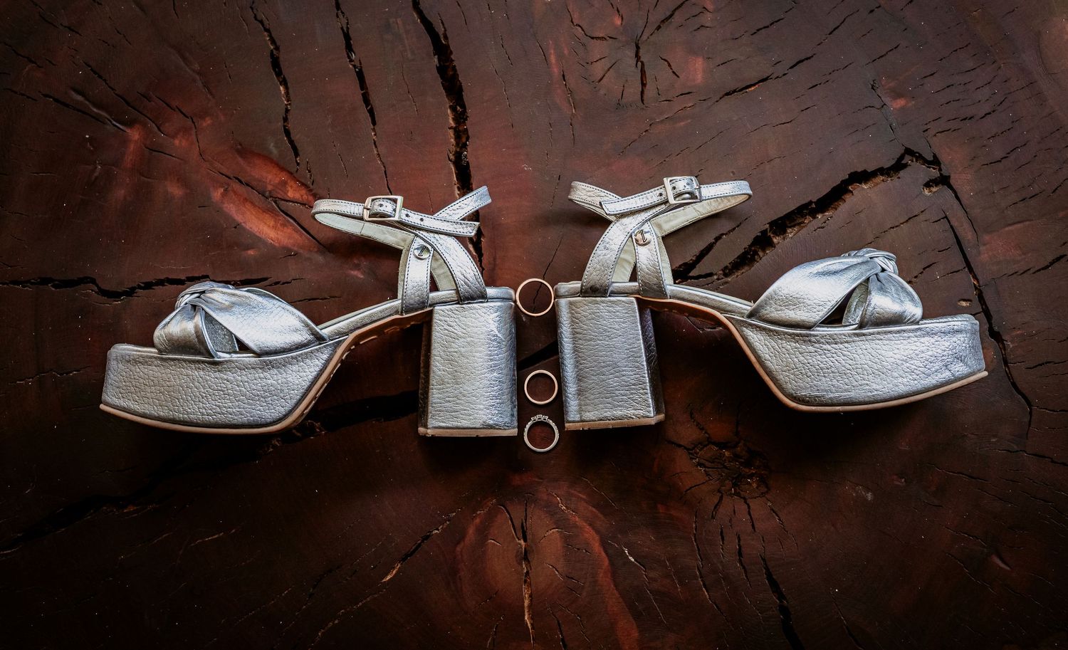 Silver platform bridal heels and two gold wedding rings displayed on a rustic wooden surface.